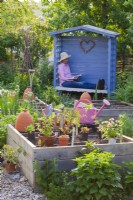Kitchen garden with seedlings and tools displayed on raised bed. In background woman in small gazebo resting from work.
