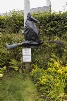 The Drunken Witch sculpture. A witch made of old wellington boots, plastic and an old fashioned witches broomstick are attched to a metal lampost with a sign saying' 'Don't drink and fly'. A variety of foliage is in the background. Harbour Lights, Devon NGS garden. July. 
