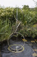 A rotating water feature, made of three interlinked hoops set in a pond. Water sprays downwards from the top hoop. A variety of water plants and other foliage is in the background. Harbour Lights, Devon NGS garden. July. 