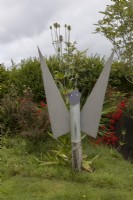 The 'Gardening Angel' made from recycled materials in front of a hedge, red monbretia, teasels and a variety of other plants with lawn in front. Harbour Lights, Devon NGS garden. July. 