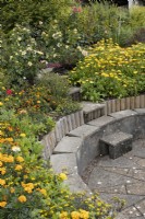 Journey to the Sun, the circular, sunken barbecue area, with steps leading down to a paved area surrounded by stone seating, amongst a profusion of yellow and orange planting. Harbour Lights, Devon NGS garden. July. 