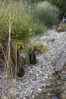 In the Bottle Garden, a variety of grasses grow within a gravel base amongst many glass bottles of various colours, with some made into sculptures including a stream. Harbour Lights, Devon NGS garden. July. 