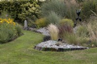 A curving lawn and path with the Bottle Garden on the right; a variety of ornamental grasses with a gravel base and a variety of glass bottles and bottle sculpture scattered within.  Harbour Lights, Devon NGS garden. July. 
