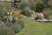 A mixed cottage style border on the left with lawn on the right and in the background a curvy bed, filled with a variety of grasses and glass bottles, as edging and sculptures. This bed is called 'The Bottle Garden'. Harbour Lights, Devon NGS garden. July. 