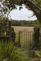 An old metal gate, partially open, leading to an open grassy field. Harbour Lights, Devon NGS garden. July. 