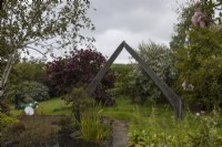 An arch, made of old wind turbine blades over a paved path. A dragonfly made of recycled materials is to the left and a variety of shruibs and trees surround the arch. Harbour Lights, Devon NGS garden. July. 
