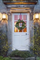 Portugese laurel and dogwood Christmas wreath hanging on house door.
Lit coach lamps and winter flowering Jasmine to either side of doorway.