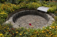 Journey to the Sun, the circular, sunken barbecue area amongst a profusion of yellow and orange planting, with a paved sun shape in the middle and seating around the edges. Harbour Lights, Devon NGS garden. July. 