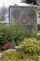 A concrete structure has a face mounted on it, made from recycled metal agricultural parts. A mix of yellow flowering plants is in the foreground with a paving slab path curving from left to right. Harbour Lights, Devon NGS garden. July. 