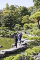 Several Japanese people walking on the stone stepping stones at the edge of the lake. 