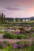 Sunrise view over the gravel garden towards Blagdon Lake - alliums, ornamental grasses, Lysimachia ciliata 'Firecracker' and mixed planting flower beds and small pond 
