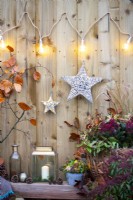 Wicker stars wrapped in lights hanging on wooden wall with strung up lights and a wooden bench with pinecones, a lantern, small metal pot with lights and Ivy and a wicker basket containing Skimmia