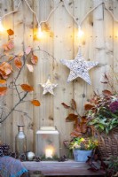 Lantern, glass bottle, bauble, pinecones, blanket and a small metal pot containing Ivy and light wrapped wicker spheres next to wicker basket containing Leucothoe on wooden bench with Beech branches behind, lights and stars hanging on the wall above