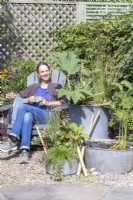 Woman sitting in garden chair behind galvanised metal containers planted with Acorus, Juncus, Gunnera, Rodgersia, Houttuynia, Isolepis , with metal basin pond planted with Cyperus and  Nymphaea with bamboo ladder and pebbles