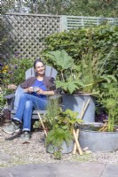 Woman sitting in garden chair behind galvanised metal containers planted with Acorus, Juncus, Gunnera, Rodgersia, Houttuynia, Isolepis , with metal basin pond planted with Cyperus and  Nymphaea with bamboo ladder and pebbles