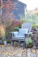 Recycled plastic chair with cushions and blanket with wicker trug containing Euphorbia, Carex and Chamaecyparis surrounded by mixed planting and leaves scattered across a wooden deck
