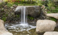 A waterfall over large stone boulders into a pond with Asplenium scolopendrium and damp loving plants growing out of a dry stone wall 