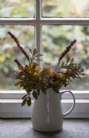 A white ceramic jug of drying flowers on a slate window sill with single glazed wooden window behind. 
