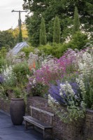 Stone walls with raised cottage style beds behind, Centranthus ruber self seeded through the border,  Wooden bench and large urn on paved patio beneath