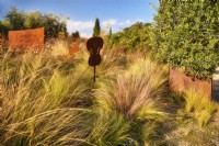 Mediterranean garden view at the entrance zone decorated with violin sculptures, grassland with Stipa tenuissima and Olive tree in the iron container in autumn time, sunlit with warm evening light.

Italy, Tuscan Maremma, Orbetello
Autumn season, October
