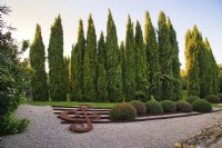 Art installation of The musical stave with treble clef sculpture and topiary bushes in the form of notes on the gravel in Mediterranean garden with rows of Cupressus Sempervirens or Italian Cypress trees on background. 
Italy, Tuscan Maremma, Orbetello
Autumn season, October
