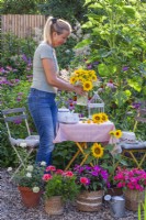 Woman placing a vase with sunflowers on the table, containers with Impatiens and Pelargonium.