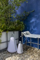 Repurposed upcycled modern contemporary metal bench seat made from an IBC - intermediate bulk container frame - blue painted rendered wall - IBC container planted with Betula pendula and Crataegus monogyna- Hawthorn - modern lights