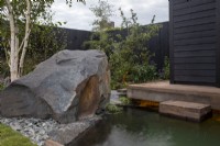 Timber frame wooden deck with steps leading down to a cold water plunge pool - a black painted fence with Malus domestica - Apple tree and Betula pendula - large stone boulder and rocks