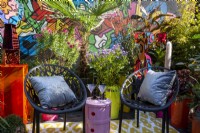 A modern contemporary colourful balcony terrace garden with black chairs and cushions - a pink table with cocktail glasses - mixed perennial container planting 