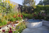 A courtyard garden with white paving slabs copper metal rill with water flowing into a raised small pool - mixed perennial planting of Echinacea purpurea, Dahlias, Kniphofia 'Papaya Popsicle' and ornamental grasses 