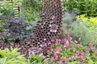 A rusty garden sculpture made from reclaimed, recycled metal materials with spaces made for an insect hotel - mixed planting of Geum 'Pink Petticoats', Foeniculum vulgare 'Purpureum', Angelica sylvestris 'Vicars Mead' - syn. 'Vicar's Mead'