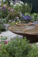 Landform Mental Wealth Garden. Designer: Nicola Hale. Reclaimed circular metal water feature with rusty hoops. Summer.