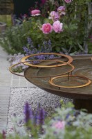 Landform Mental Wealth Garden. Designer: Nicola Hale. Reclaimed circular metal water feature with rusty hoops. Plants include rose, salvias and herbs. Summer.