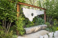 An oak bench seat with a contemporary sculpture made from reclaimed salvaged concrete suspended from rusty steel girders - mixed perennial planting with Corylus avellana - Hazel trees either side