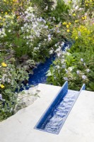 A modern contemporary water rill made from recycled plastics set in stone mixed perennial planting of Geraniums, Lychnis flos-cuculi 'White Robin' and Ranunculus acris 