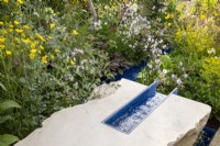 A modern contemporary water rill made from recycled plastics set in stone, mixed perennial planting of Euphorbia robbiae, Geranium, Lychnis flos-cuculi 'White Robin' and Ranunculus acris 