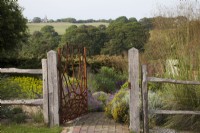 Drought tolerant garden filled with mediterranean plants. Bespoke Steel Gate allowed to rust, depicting corn and the fields, framing the view of the fields to Pett Church and entrance to The Jewel Garden -  Curved brick pathway. Nepeta 'Six Hills Giant' - Catmint,   Santolina chamaecyparissus ' Yellow Buttons', Euphorbia seguieriana subsp. niciciana , grasses including Calamagrostis brachytricha and Stipa gigantea, Geraniums. Gate Made by  Jake Bowers The Thirsty Bear Forge Hastings
