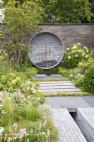 Garden wall made from stacked concrete slabs, a circular water feature with salvaged brass taps, mixed perennial planting in white and green colours, grey clay brick pavers and stone paving