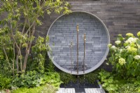 Garden wall with a circular concrete and clay brick pavers water feature, brass tap spouts and copper pipes, Hydrangea arborescens 'Annabelle'