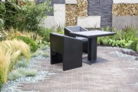 Brick paved patio area with black table and chairs, wall made from grey brick and concrete slabs - mixed perennial planting 