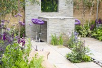 Stone paved patio with water rill, dining area with purple bar stools, mixed perennial planting in purple and white colours