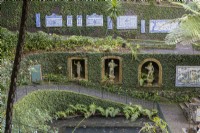Several paths cross the steep sides of a valley with retaining walls, covered in ivy and with paths in front. On the bottom path, arched stone windows create niches with stone figurative statues in. A path above has antique tiled panels set on it. Monte Palace Gardens, Madeira. August. 