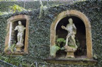 Old stone window arches set into ivy covered walls create niches for stone figurative statues. Monte Palace Gardens, madeira. August. 