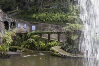 Looking out from behind the  waterfall on to the Central Lake, with spouts of water from the Fortress on the left and various paths sloping and curving in the background. Monte Palace Gardens, Madeira. August. Summer