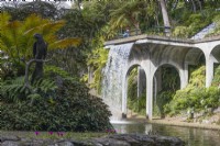 The waterfall, designed by Jose Berardo, flows from a viaduct style balcony into the Central Lake. A bird statue is on an island with tropical planting in the foreground. Monte Palace Gardens, Madeira. August. Summer
