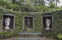 A curved ivy covered wall, with three stone window arches inset in the wall and figurative statues within the niches. A bench with the hashtag MontePalaceGardens is in the foreground. Monte Palace gardens, Madeira. August. 