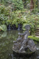 A figurative statue in a pond, with small waterfalls in the background, steps to the right and lush tropical planting in the background. Monte Palace Gardens, Madeira