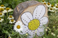 A tree stump made into an insect hotel with a painted daisy flower for decoration, planting of Echinacea purpurea 'White Swan'