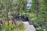 Steps leading to black metal infinity pool overlooked by a large sculptured face in 'The Thrive Reflective Mind Garden' - RHS Chatsworth Flower Show 2019, June