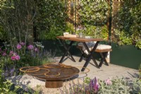 An urban courtyard garden with wooden bench and table, corten steel water feature, raised bed containers planted with Trachelospermum jasminoides climbing over hazel fencing screens